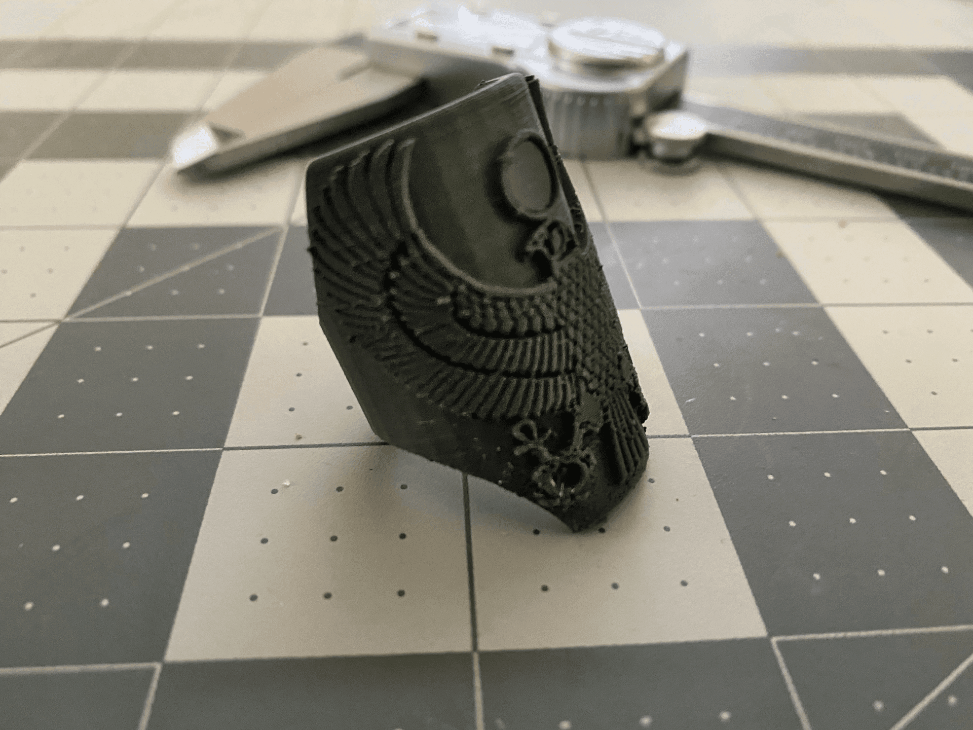 Black 3D-printed object with intricate design on checkered background near measuring tool.